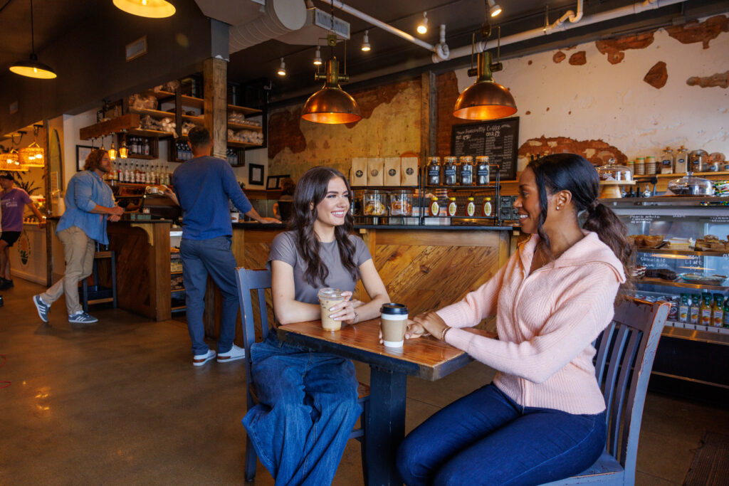 Women sitting at table with coffee while two men stand in background at coffee shop