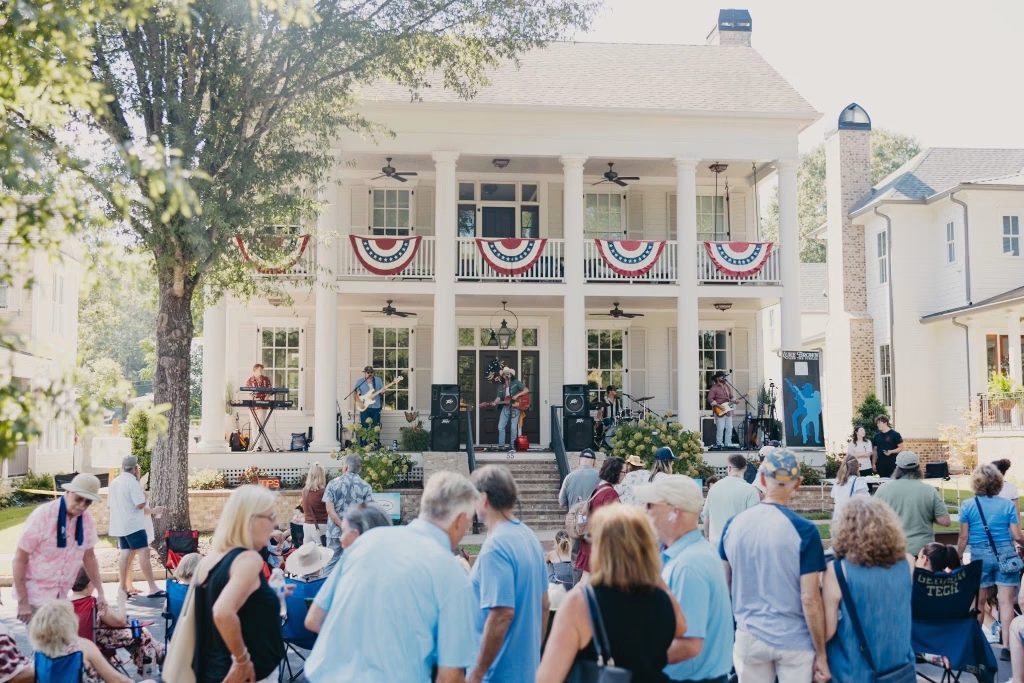 Exterior view of people gathered on the front lawn of a historic home, enjoying live music.
