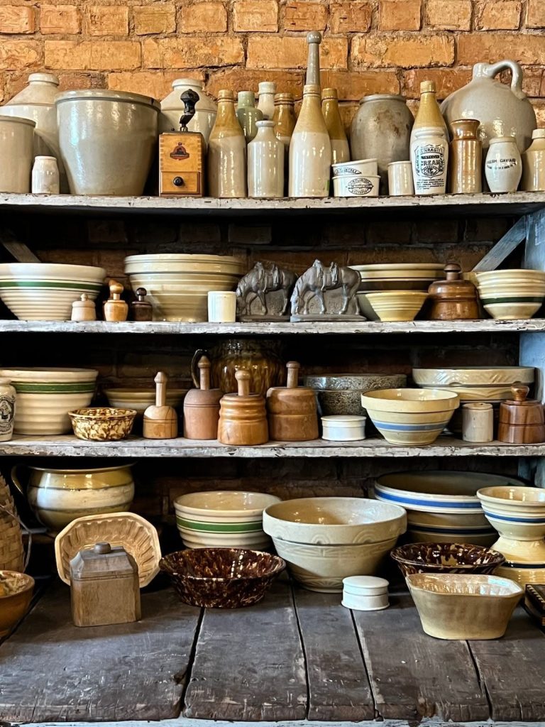 A rustic display of various vintage ceramic bowls and stoneware pottery arranged on wooden shelves against a brick wall.
